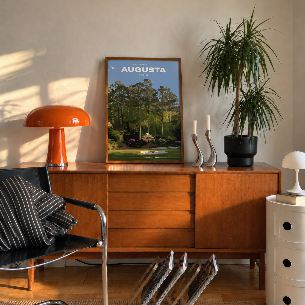 Augusta 12th hole golf print of a par 3 green below azaleas and pines, framed on a mid-century sideboard beside an orange lamp