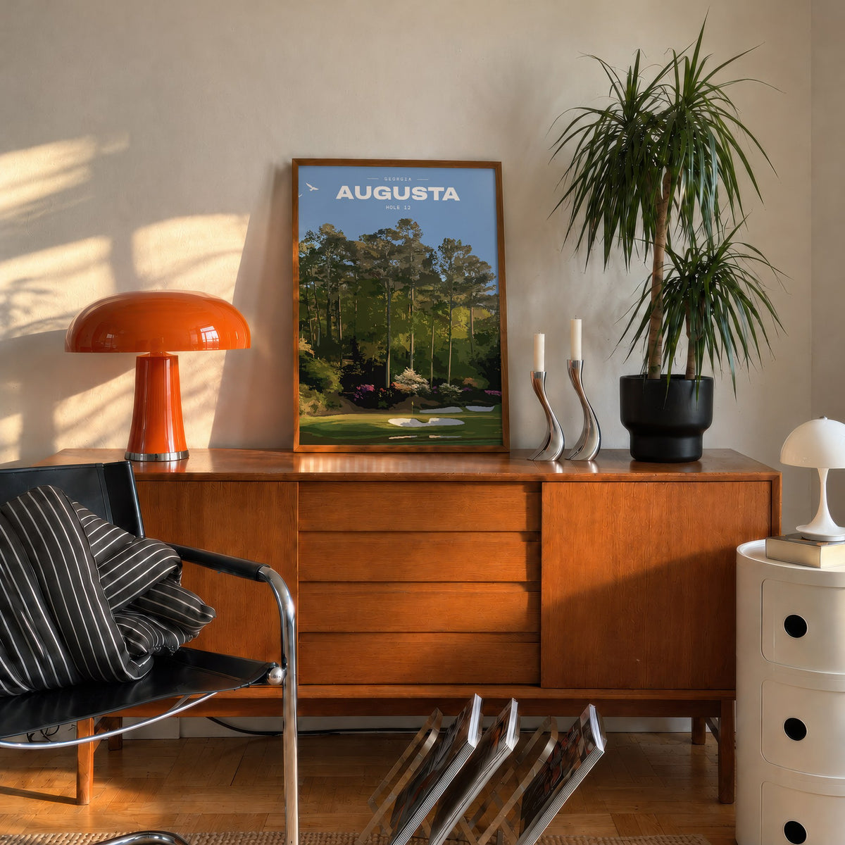 Augusta 12th hole golf print of a par 3 green below azaleas and pines, framed on a mid-century sideboard beside an orange lamp