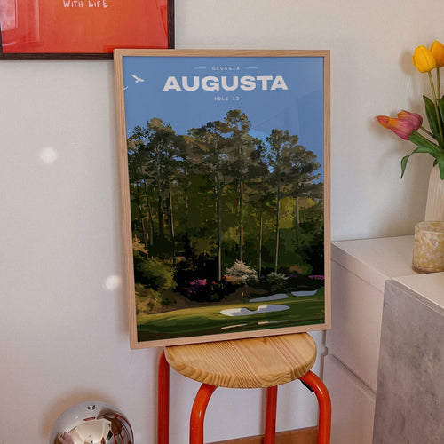 Augusta 12th hole golf print of a par 3 green below azaleas and pines, framed on an orange stool beside tulips