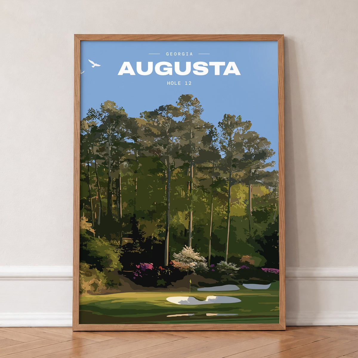 Augusta 12th hole golf print of a par 3 green below azaleas and pines, in an oak frame, leaning against a wall on a herringbone floor