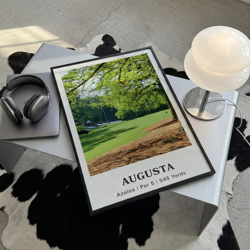 Augusta 13th hole golf print of a par 5 fairway under oak and pines, framed, lying on a steel table on a cowhide rug