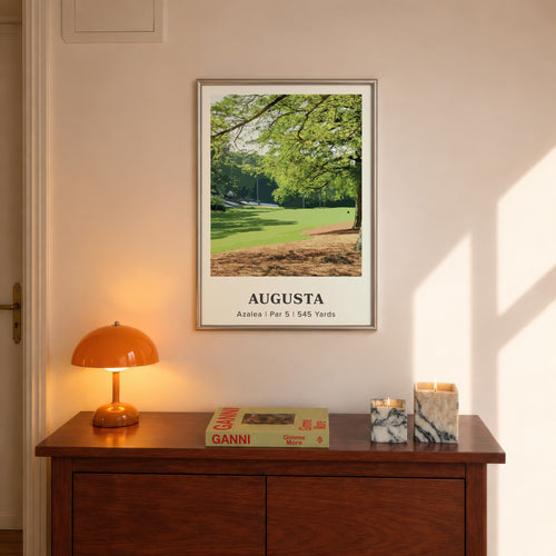Augusta 13th hole golf print of a par 5 fairway under oak and pines, framed above a console beside an orange lamp