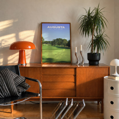 Augusta 1st hole golf print of an uphill fairway to a yellow flag, framed on a mid-century sideboard beside an orange lamp
