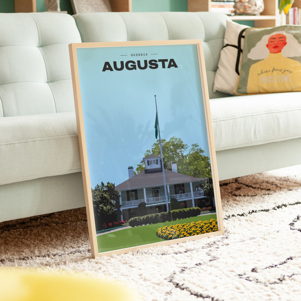 Augusta The Clubhouse golf print of the white clubhouse and yellow flowers, framed, leaning against a sofa