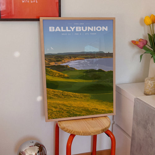 Ballybunion 11th hole golf print of dunes running down to an Atlantic bay, framed on an orange stool beside tulips