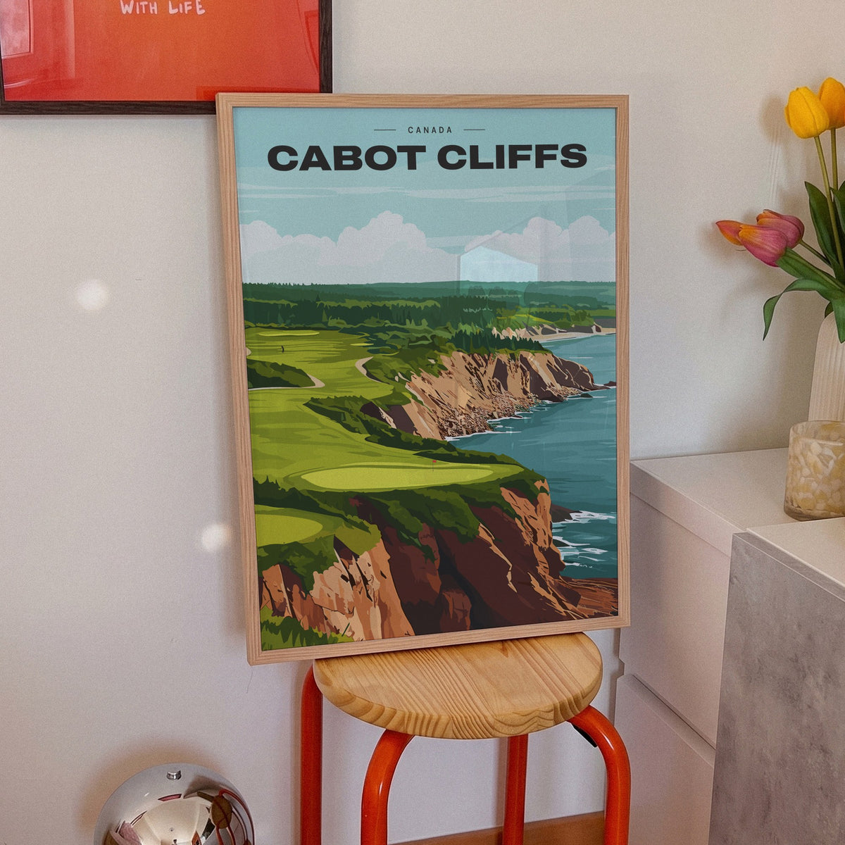 Cabot Cliffs golf print of a green on red clifftops above the sea, framed on an orange stool beside tulips