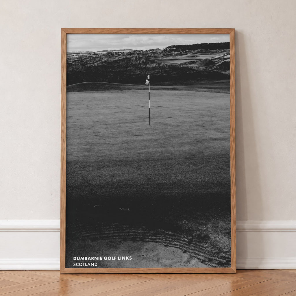 Dumbarnie golf print of a black and white photo of a flag below dunes, in an oak frame, leaning against a wall on a herringbone floor