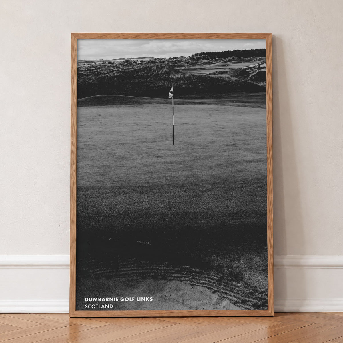 Dumbarnie golf print of a black and white photo of a flag below dunes, in an oak frame, leaning against a wall on a herringbone floor