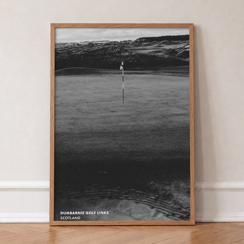 Dumbarnie golf print of a black and white photo of a flag below dunes, in an oak frame, leaning against a wall on a herringbone floor