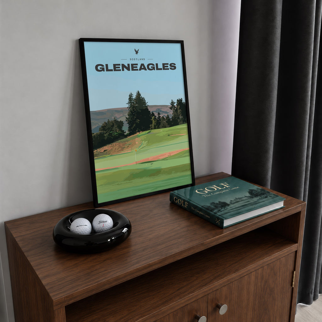 Gleneagles golf print of a green below pines and Perthshire hills, framed on a walnut cabinet beside golf balls and a golf book