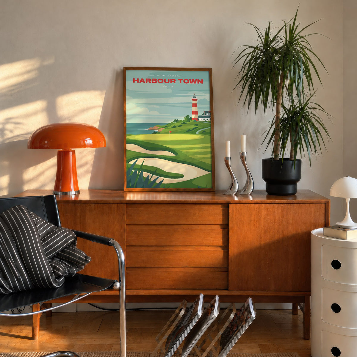 Harbour Town 18th hole golf print of a green below a striped lighthouse, framed on a mid-century sideboard beside an orange lamp