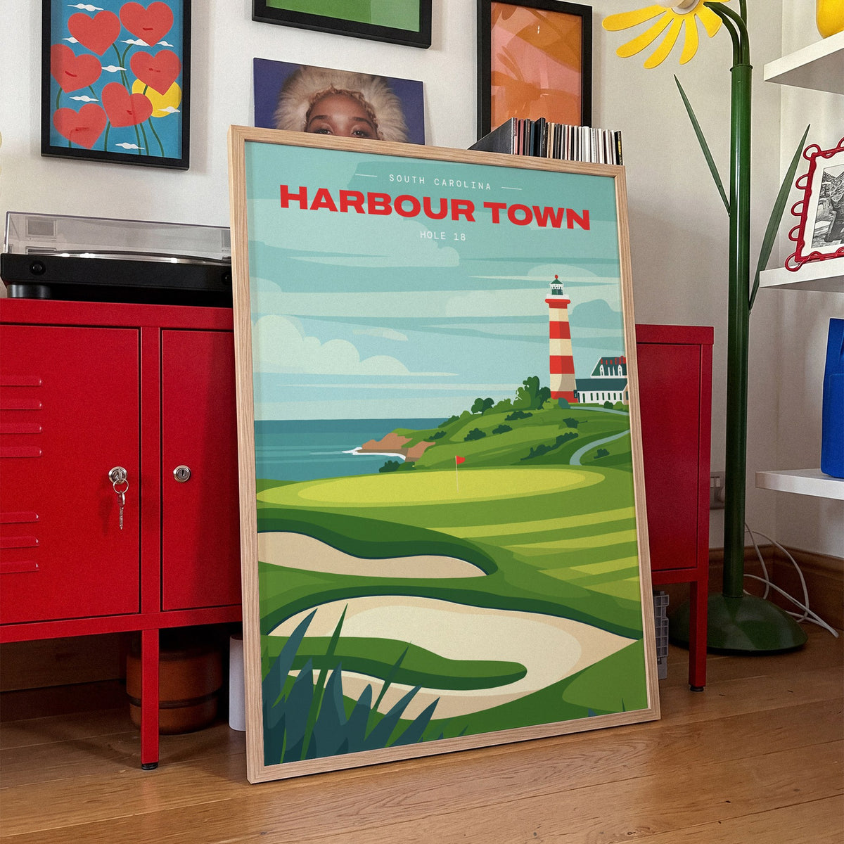 Harbour Town 18th hole golf print of a green below a striped lighthouse, framed, leaning against red lockers
