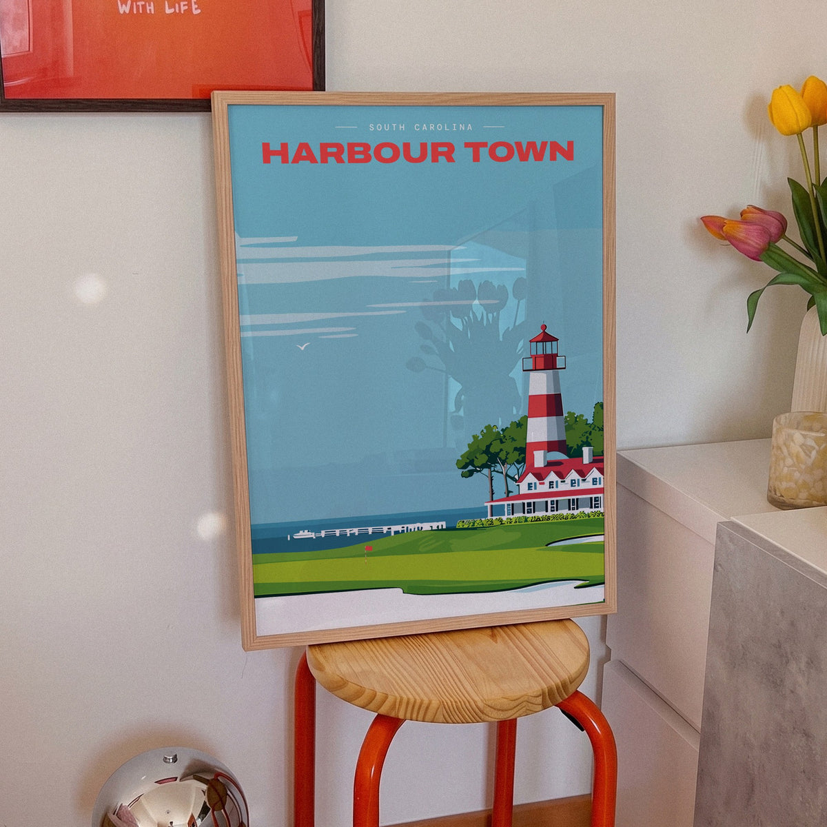 Harbour Town Lighthouse golf print of a striped lighthouse above a marina, framed on an orange stool beside tulips