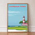 Harbour Town Lighthouse golf print of a striped lighthouse above a marina, in an oak frame, leaning against a wall on a herringbone floor