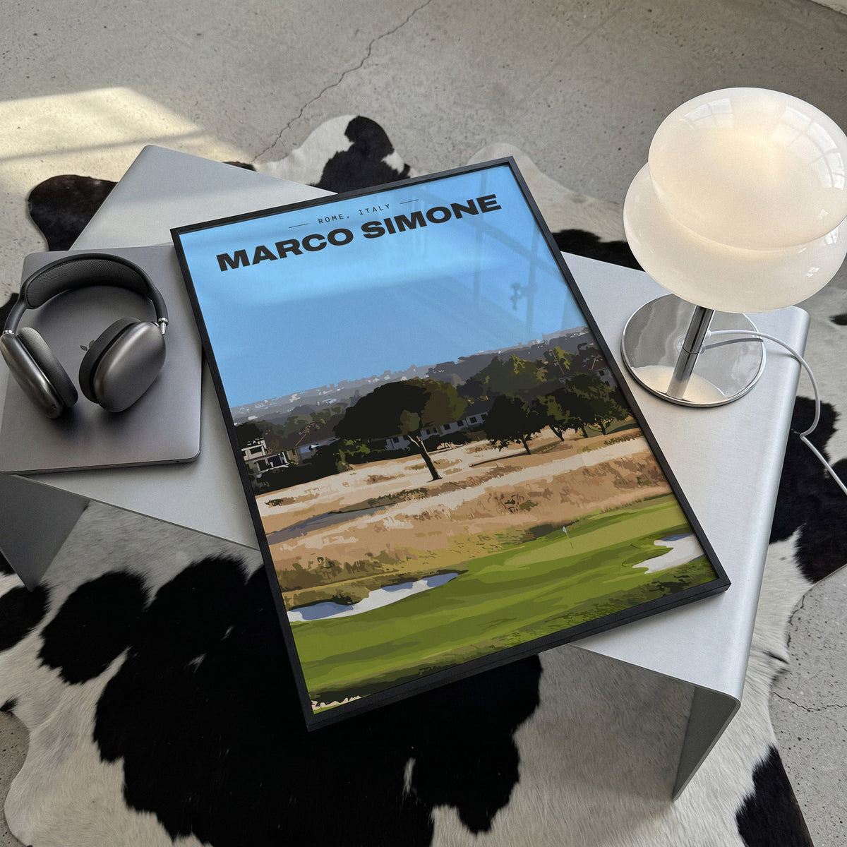 Marco Simone golf print of a green below umbrella pines and gold rough, framed, lying on a steel table on a cowhide rug