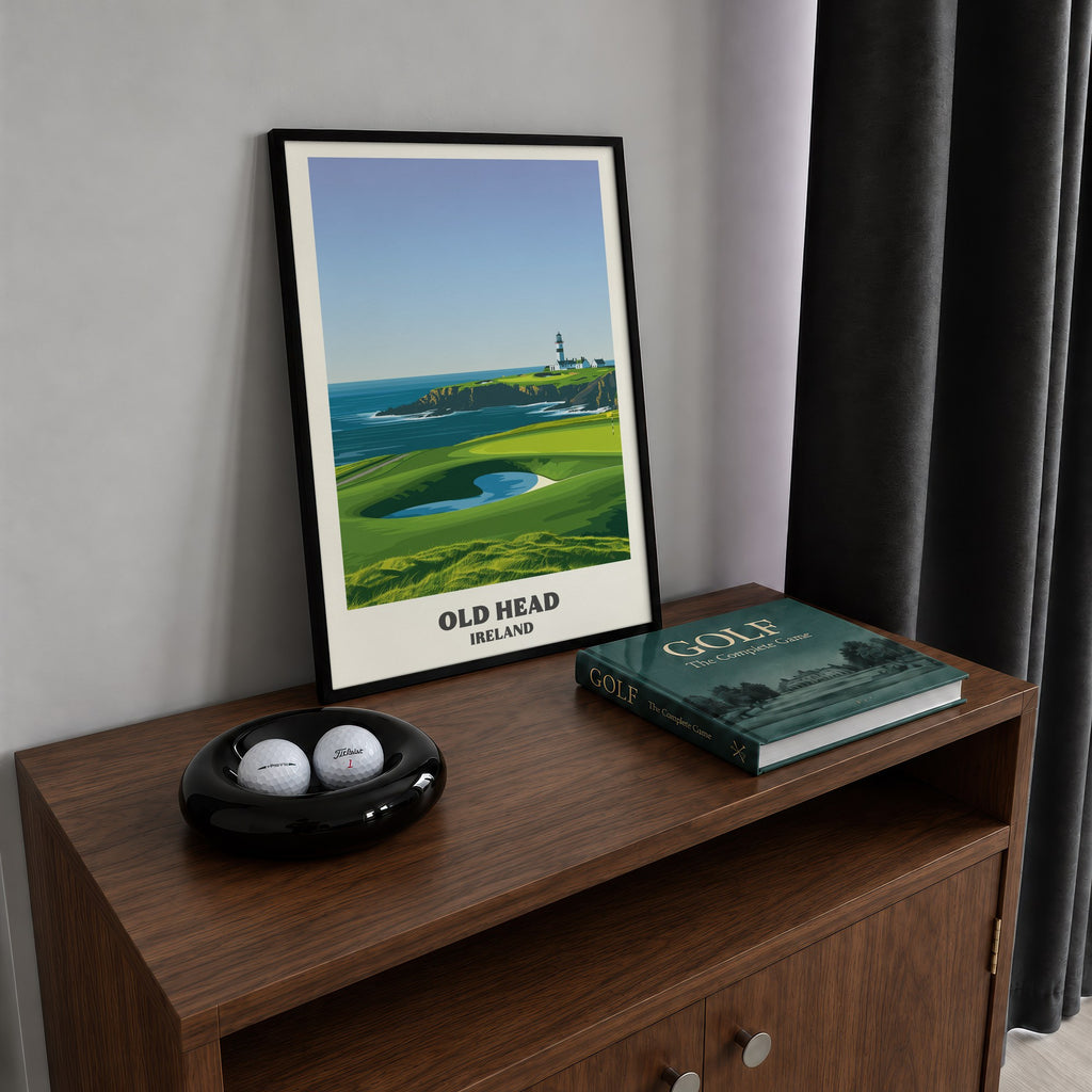 Old Head golf print of a lighthouse on a headland above a green, framed on a walnut cabinet beside golf balls and a golf book