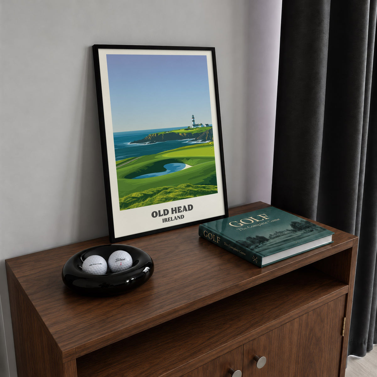 Old Head golf print of a lighthouse on a headland above a green, framed on a walnut cabinet beside golf balls and a golf book