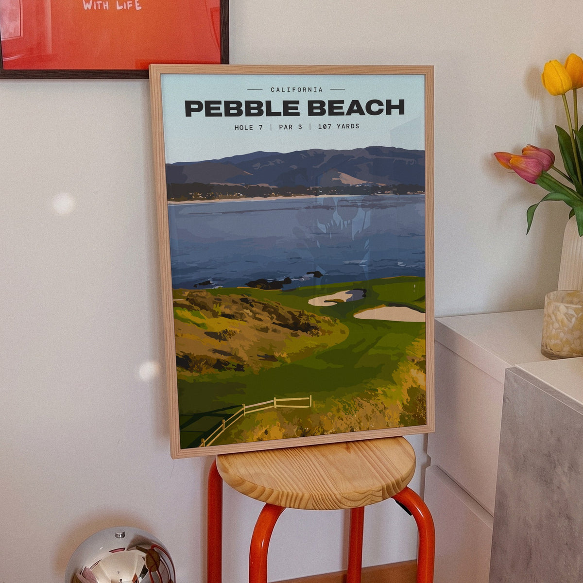 Pebble Beach 7th hole golf print of a small green on a point above the bay, framed on an orange stool beside tulips