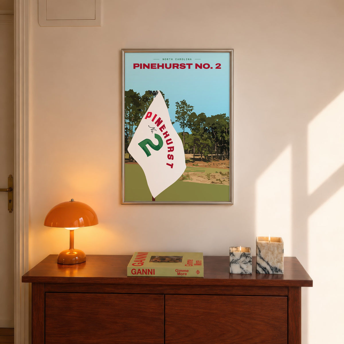Pinehurst No. 2 golf print of a white pin flag before sandy waste and pines, framed above a console beside an orange lamp