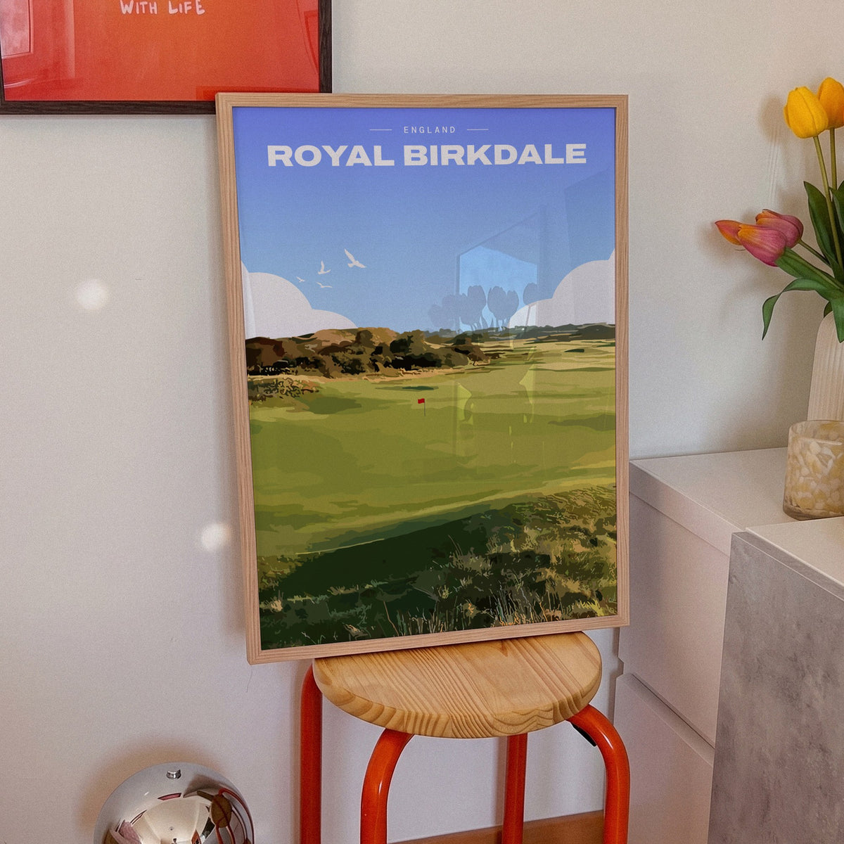 Royal Birkdale golf print of a links fairway and red flag between dunes, framed on an orange stool beside tulips