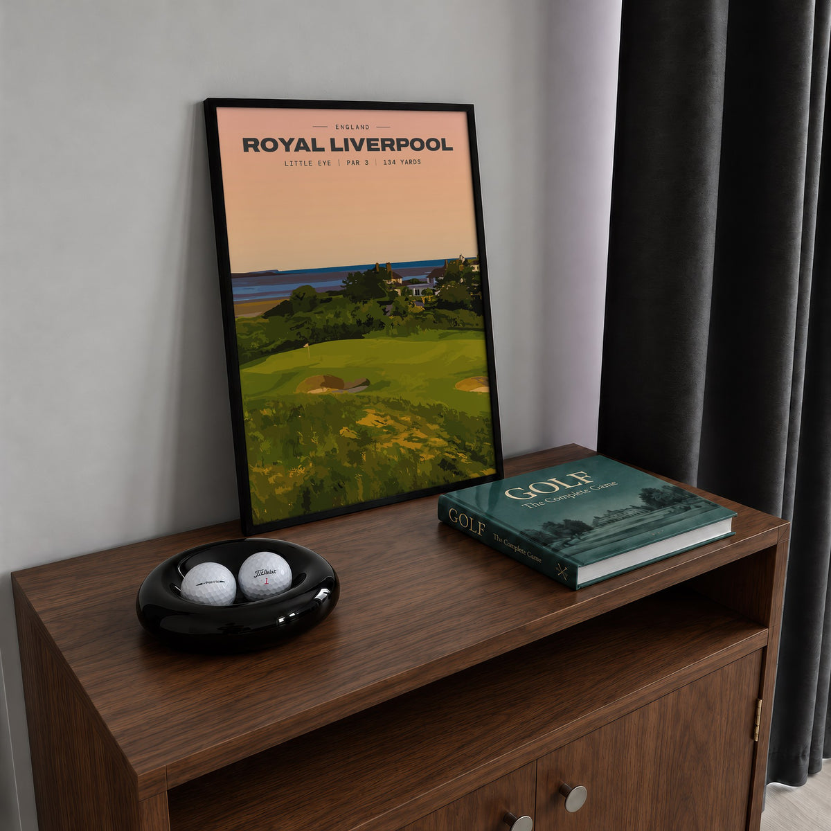 Royal Liverpool Little Eye golf print of a raised par 3 green at sunset, framed on a walnut cabinet beside golf balls and a golf book