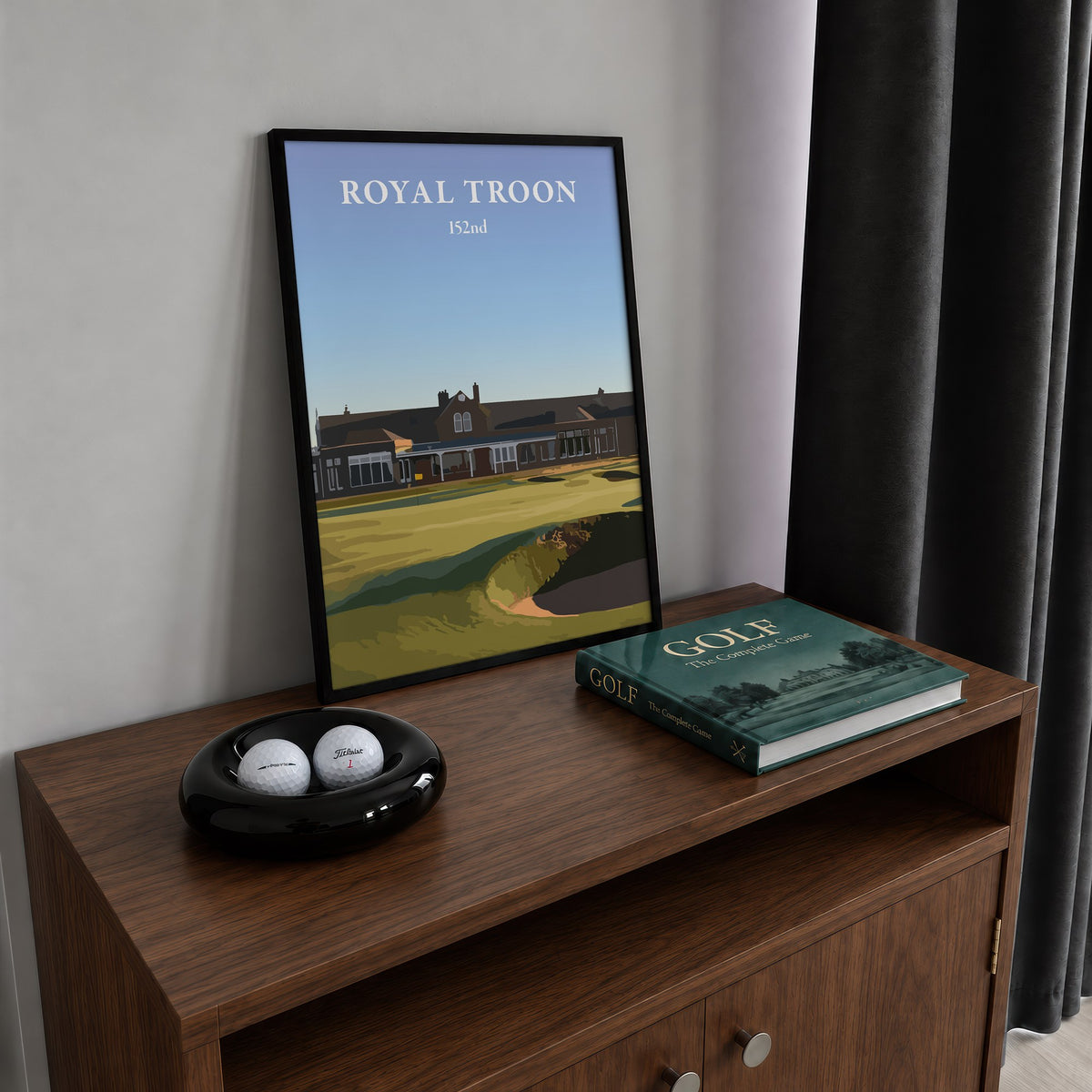 Royal Troon 2024 golf print of the clubhouse behind the 18th green, framed on a walnut cabinet beside golf balls and a golf book