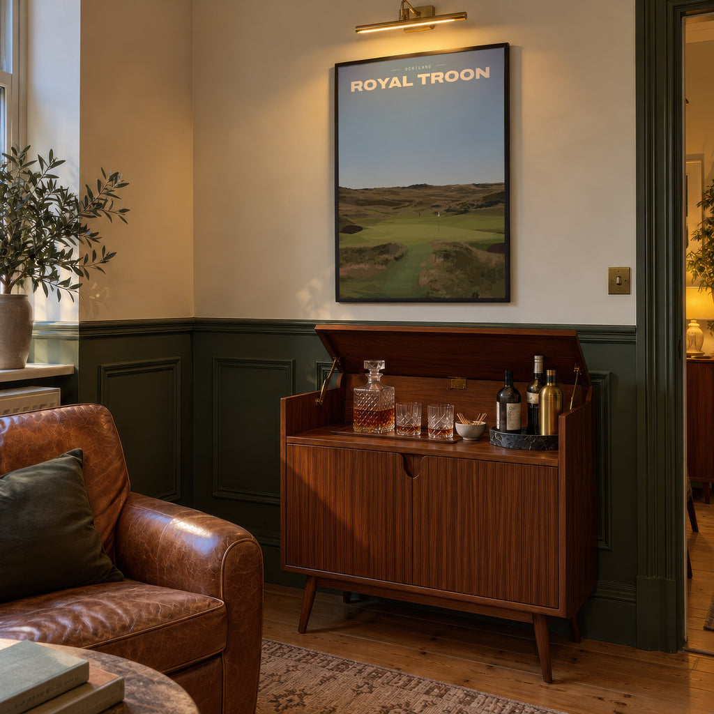 Royal Troon golf print of a links fairway through dunes to a flag, framed above a bar cabinet beside a leather armchair