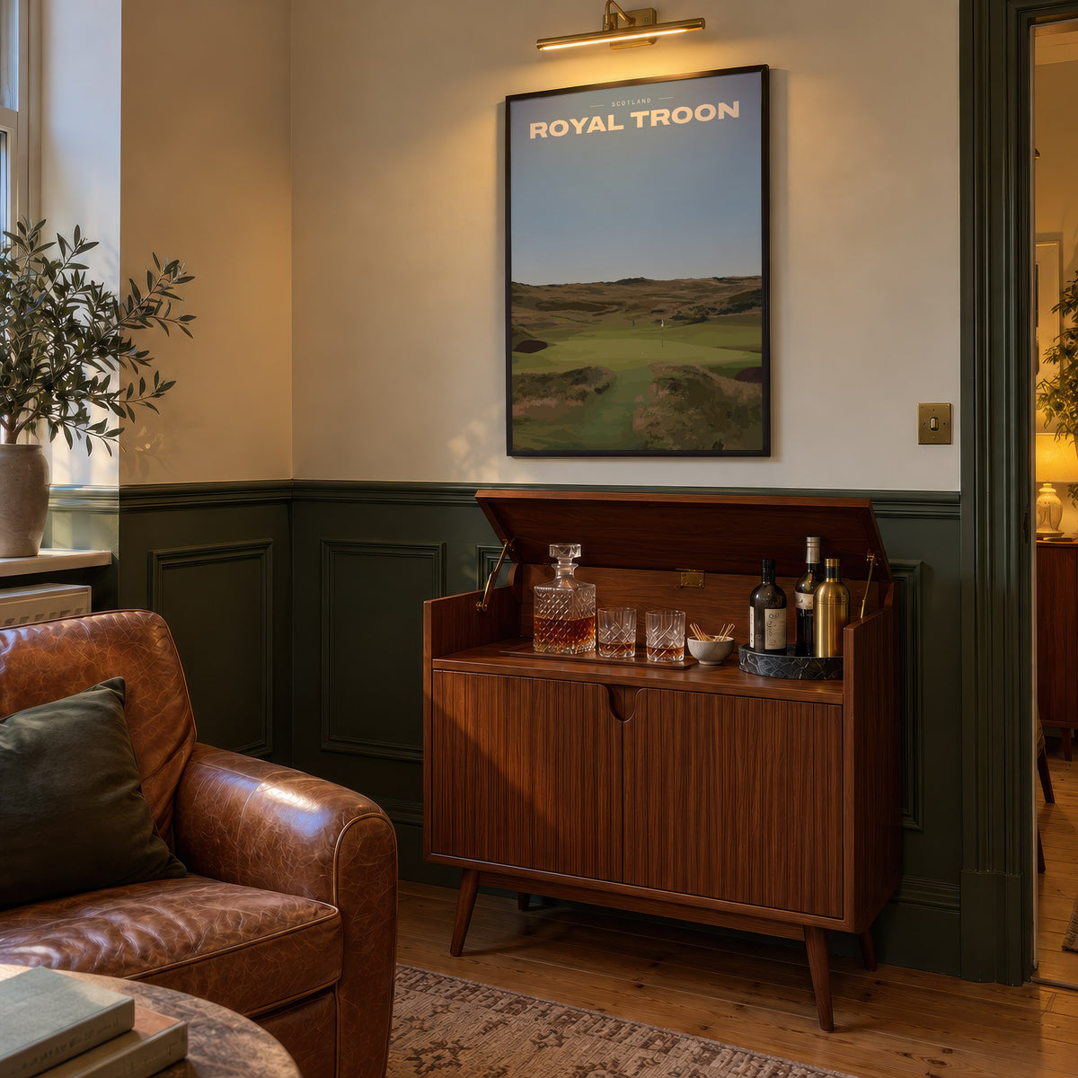 Royal Troon golf print of a links fairway through dunes to a flag, framed above a bar cabinet beside a leather armchair