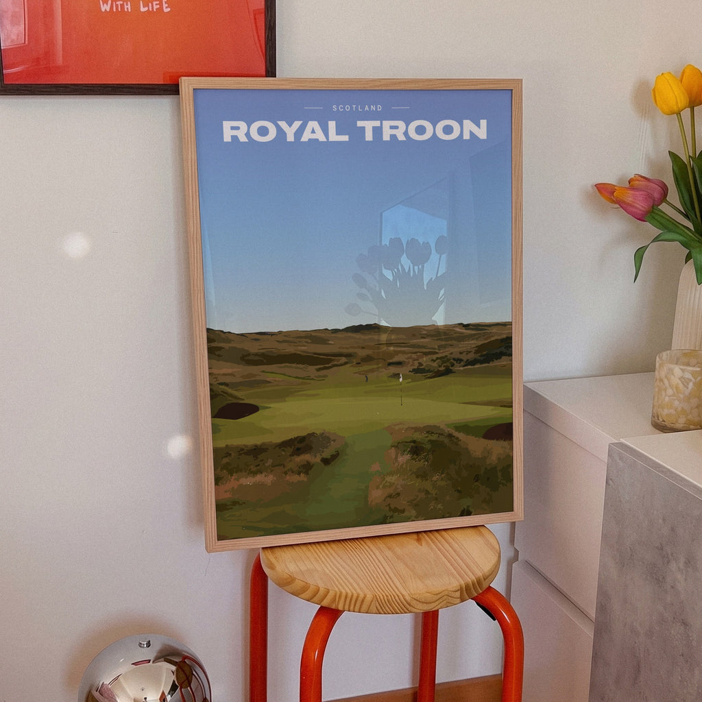 Royal Troon golf print of a links fairway through dunes to a flag, framed on an orange stool beside tulips