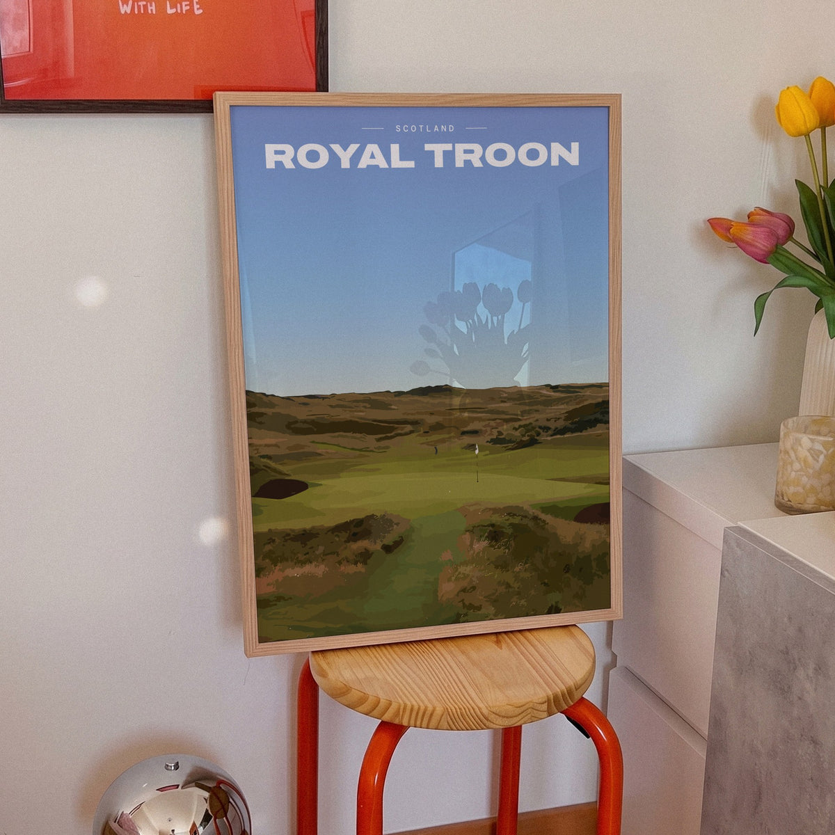 Royal Troon golf print of a links fairway through dunes to a flag, framed on an orange stool beside tulips