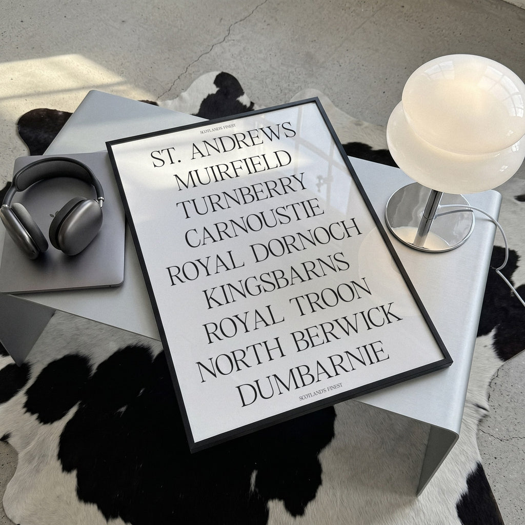 Scotland's Finest golf print of nine Scottish course names in serif type, framed, lying on a steel table on a cowhide rug