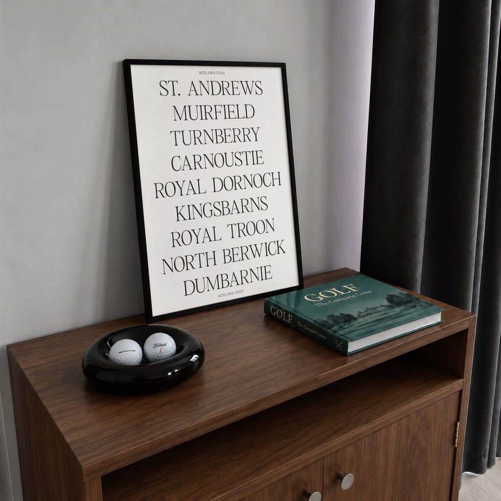 Scotland's Finest golf print of nine Scottish course names in serif type, framed on a walnut cabinet beside golf balls and a golf book