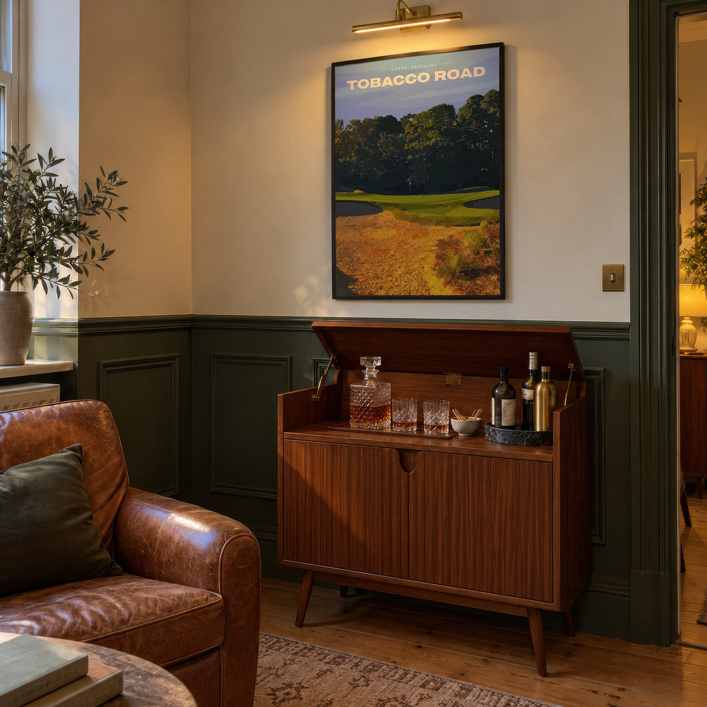 Tobacco Road golf print of a green behind golden sand and pines, framed above a bar cabinet beside a leather armchair