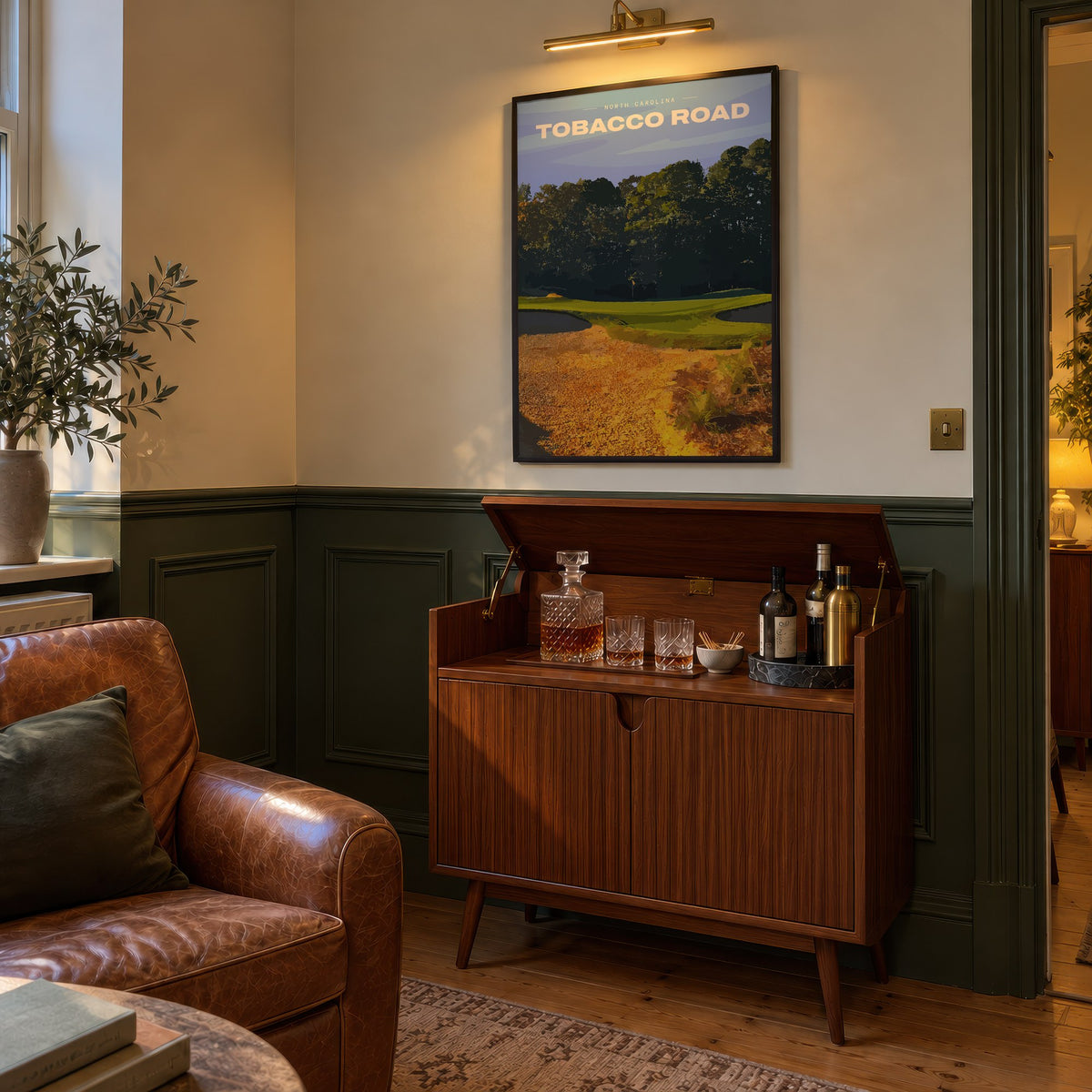 Tobacco Road golf print of a green behind golden sand and pines, framed above a bar cabinet beside a leather armchair