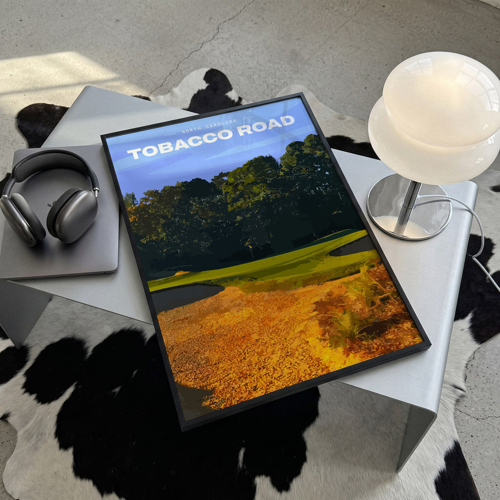 Tobacco Road golf print of a green behind golden sand and pines, framed, lying on a steel table on a cowhide rug