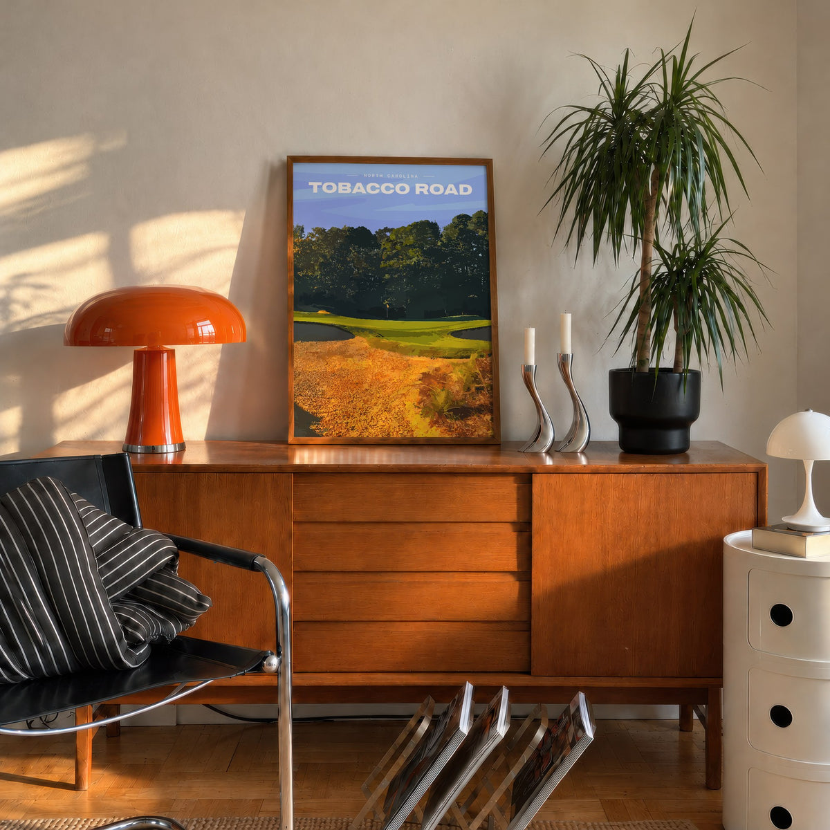 Tobacco Road golf print of a green behind golden sand and pines, framed on a mid-century sideboard beside an orange lamp
