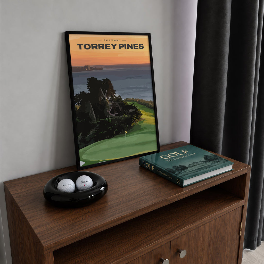 Torrey Pines golf print of a clifftop green above the Pacific at sunset, framed on a walnut cabinet beside golf balls and a golf book