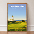 Turnberry 9th hole golf print of a white lighthouse above a links green, in an oak frame, leaning against a wall on a herringbone floor
